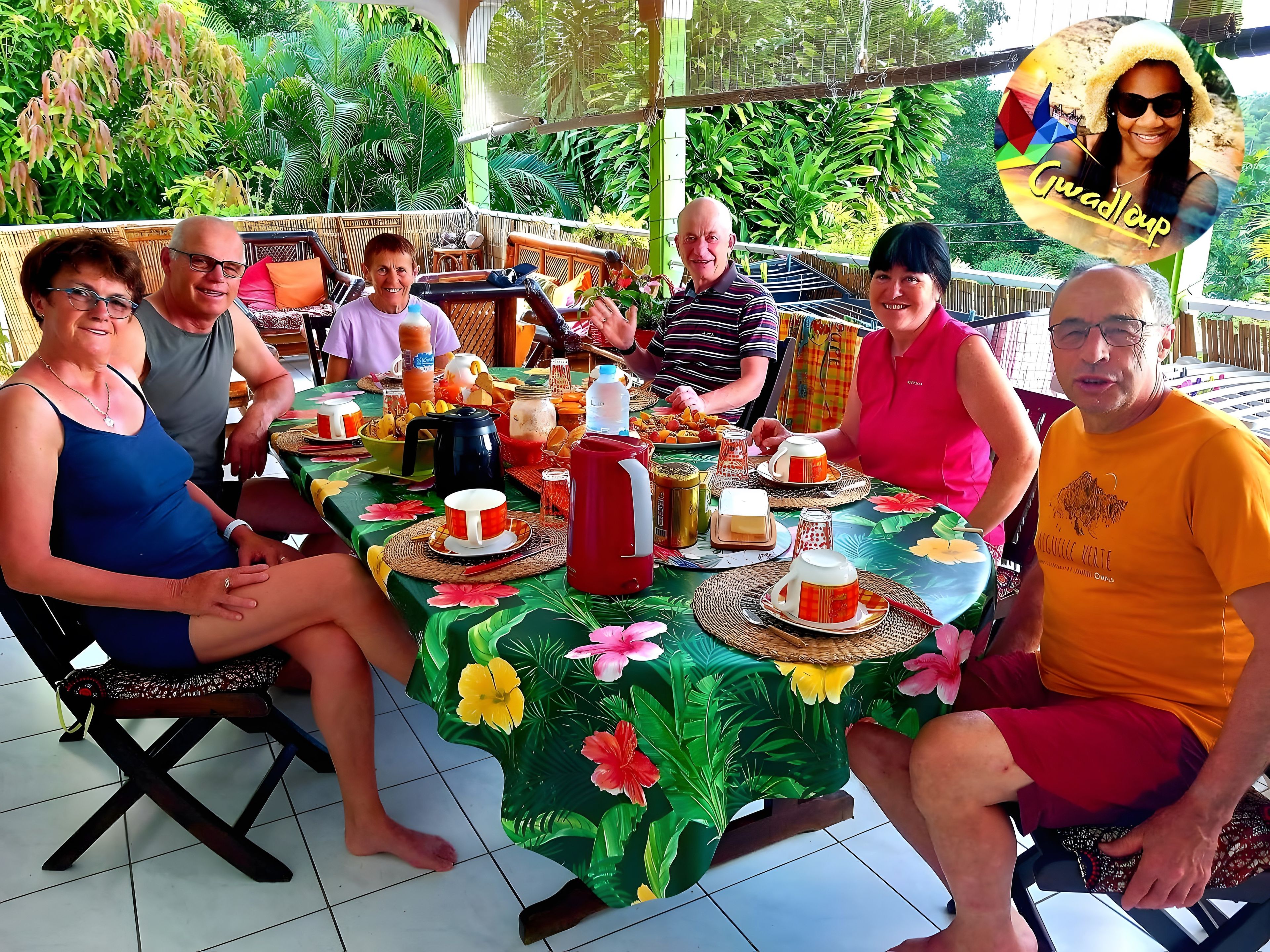 A group of people seated around a table with various food items and drinks, in an outdoor setting with lush greenery in the background.