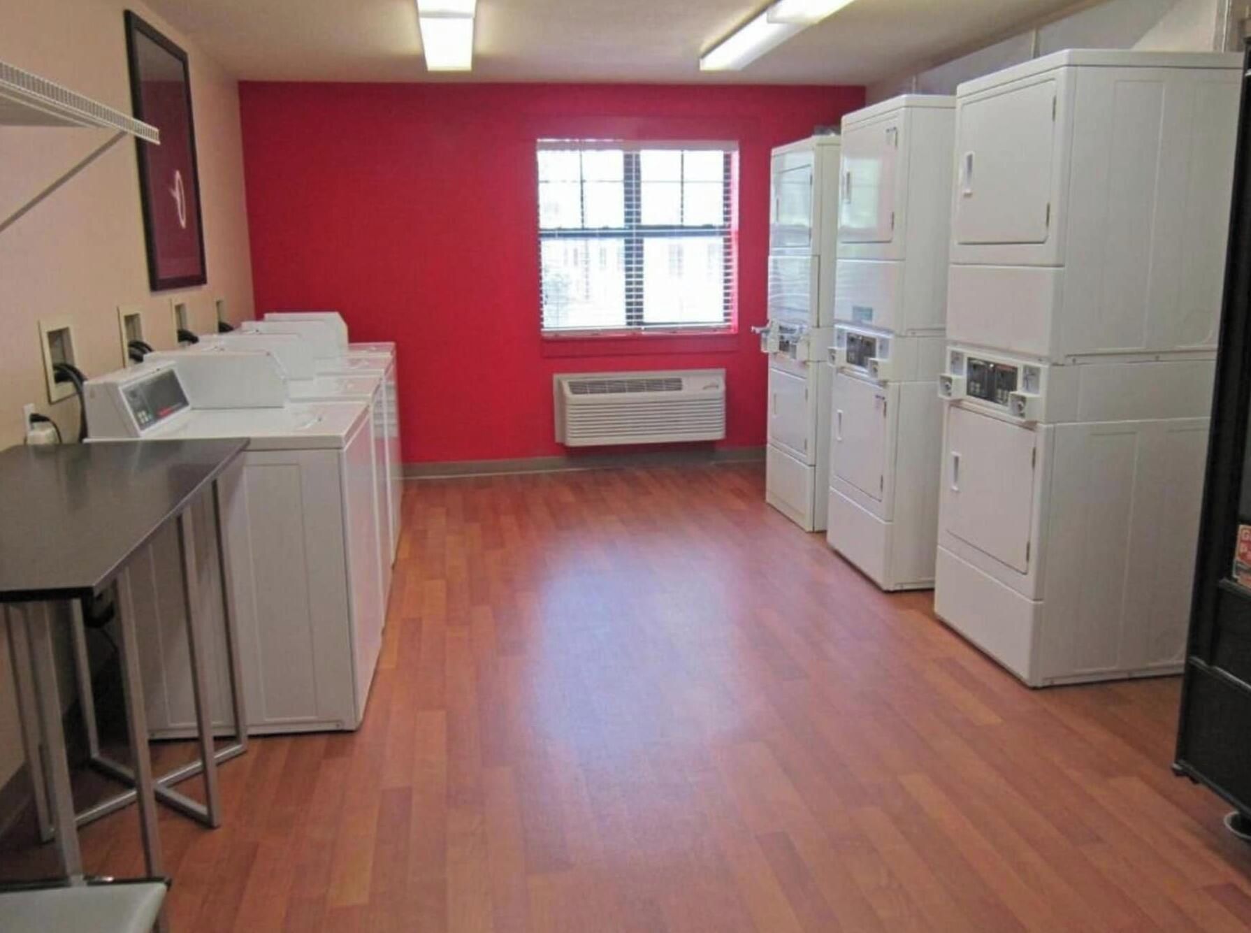 A laundry room with multiple washing machines, a window with blinds, and a red wall.