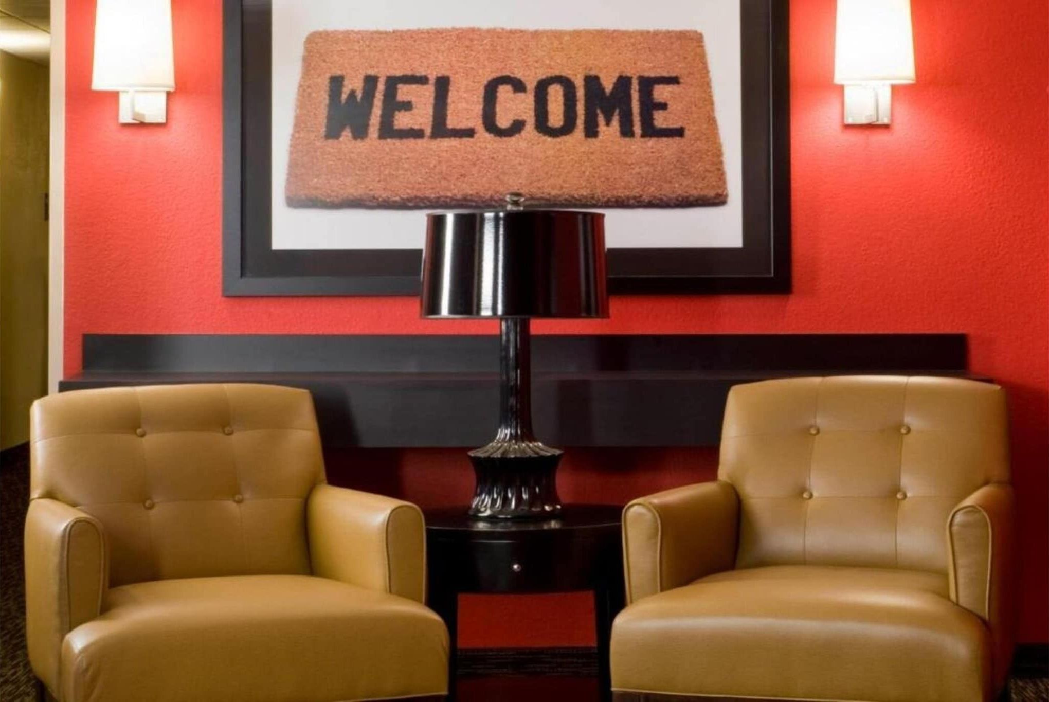 Een hotel lobby met een rode muur, een welkomstmat, twee leren fauteuils en een bijzettafeltje met een lamp.