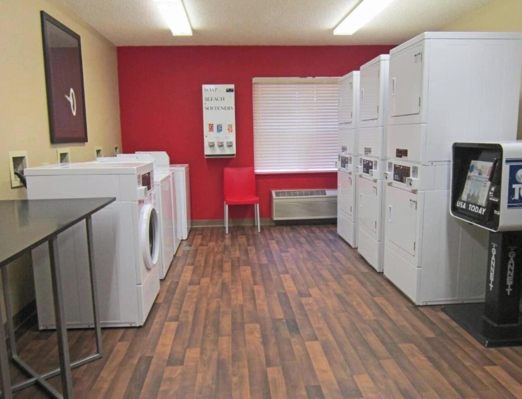 A laundry facility with multiple washing machines, a red wall, and a USA Today newspaper dispenser.