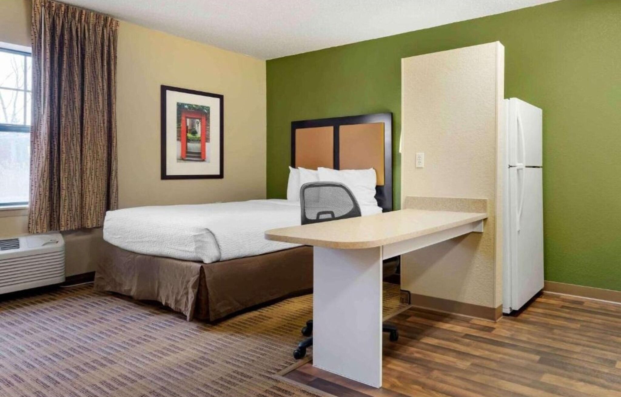 A hotel room with a bed, a desk, a chair, a refrigerator, and a window with curtains.
