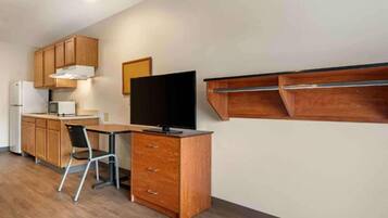 A compact kitchen with wooden cabinets, a microwave, a refrigerator, a flat-screen TV, a foldable chair, and a wooden shelf unit.