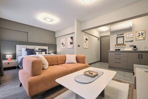 A modern hotel room with a sofa, a coffee table, a bed with a headboard, a bedside table with a lamp, and a kitchenette area. - Modern 2BR Living Areas, 2 Baths, Kadıköy, (Istanbul)