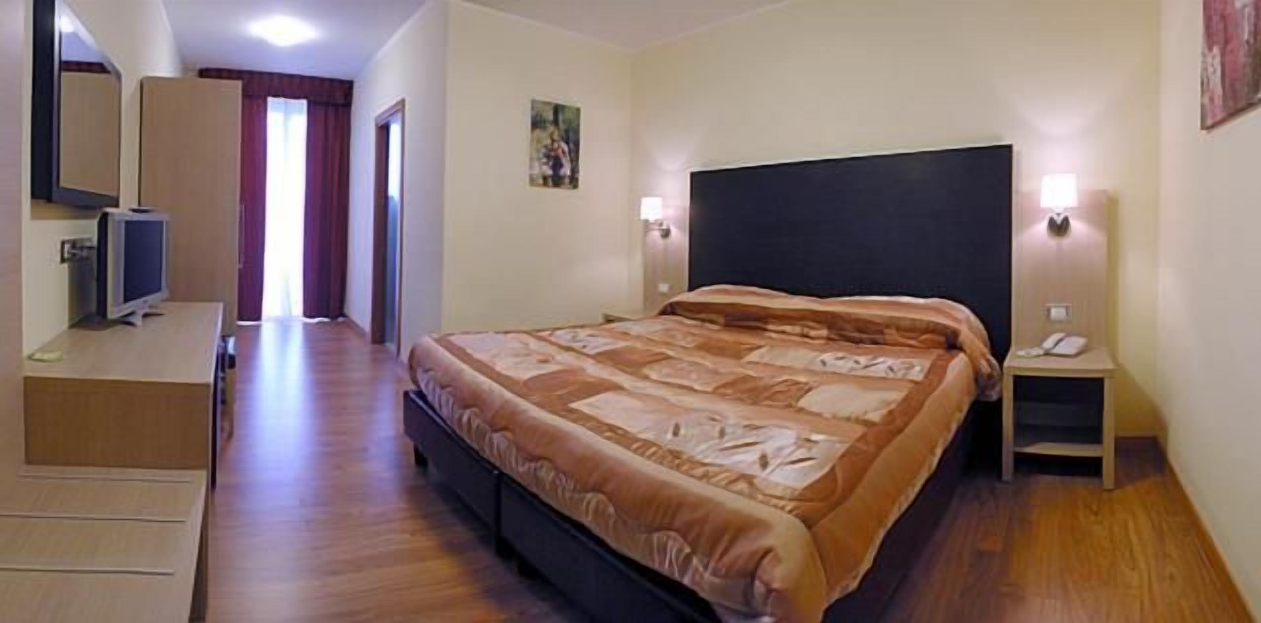 Quarto de hotel com cama, luminárias laterais, escrivaninha com computador e televisão.