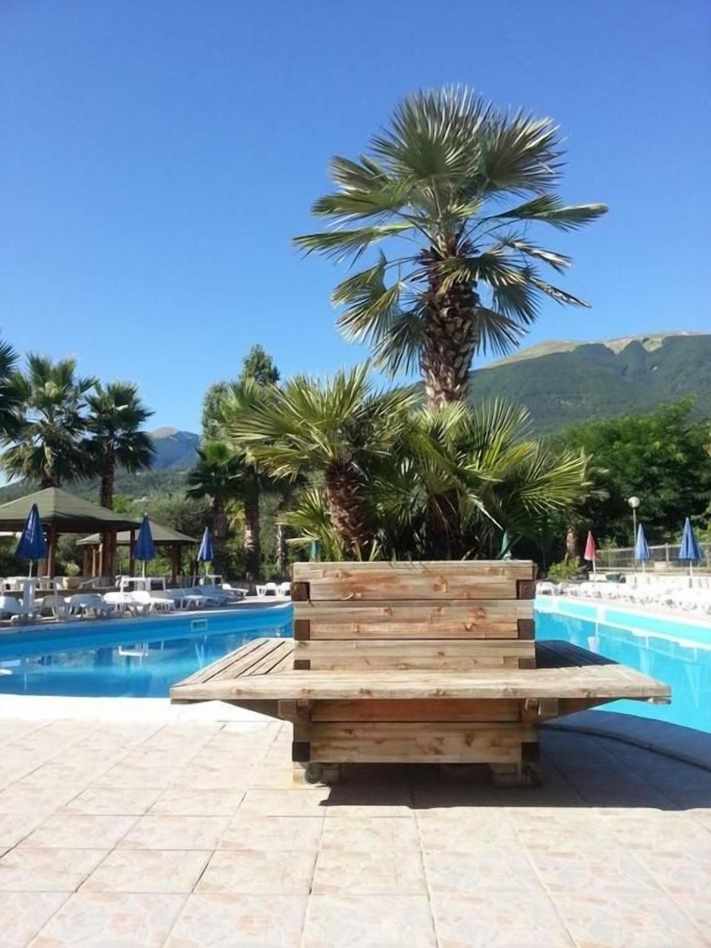 Um banco de madeira junto a uma piscina, com palmeiras e uma montanha ao fundo.