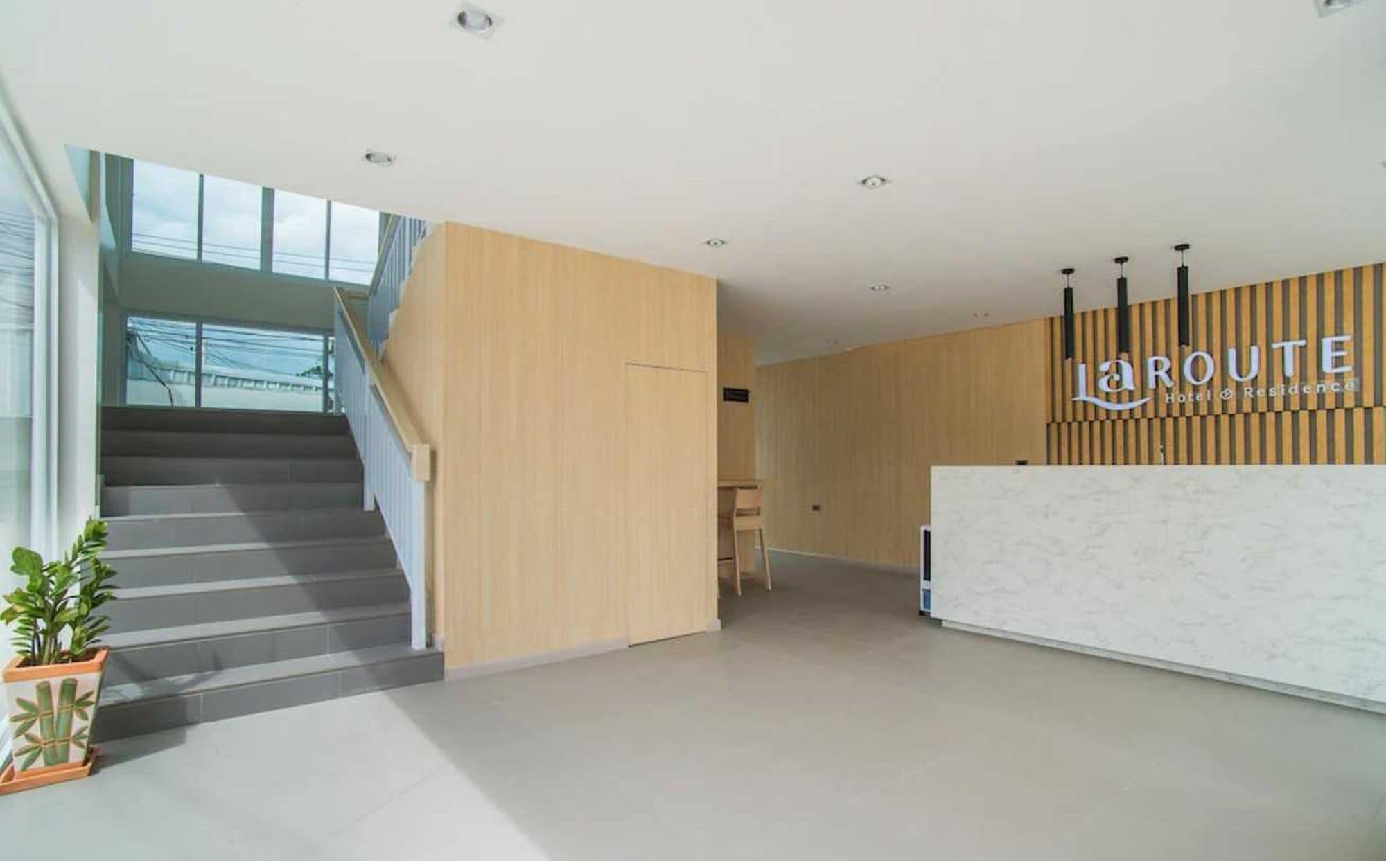 A hotel reception area with a staircase leading to an upper floor, a reception desk, and a sign that reads 'LAROUTE Hotel & Residence'.