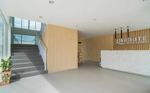 Zona de recepción de un hotel con una escalera que conduce a un piso superior, un mostrador de recepción y un letrero que dice 'LAROUTE Hotel & Residence'.