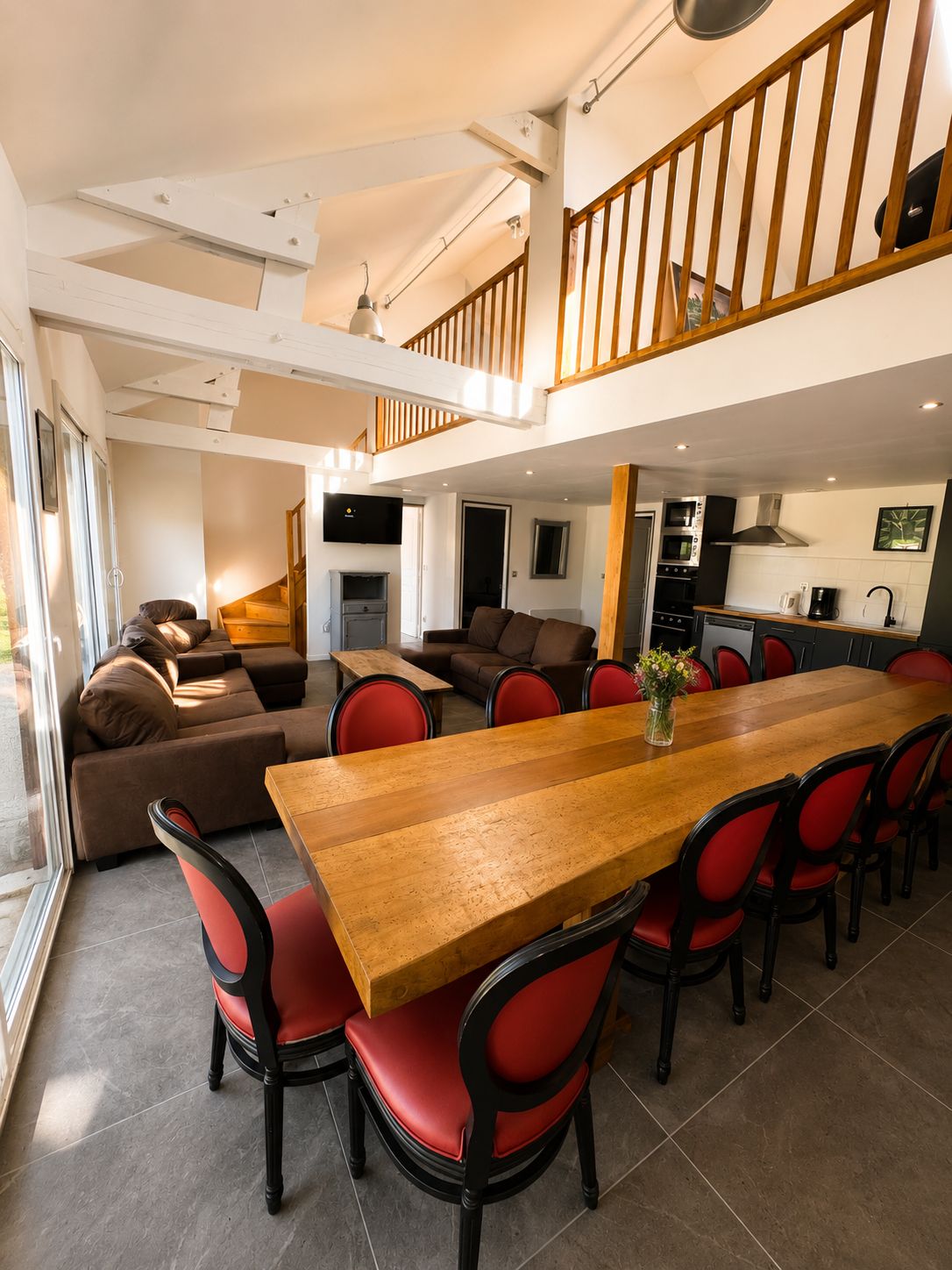 Een ruime woonkamer met een houten eettafel, rode stoelen, een bank en een keuken op de achtergrond.