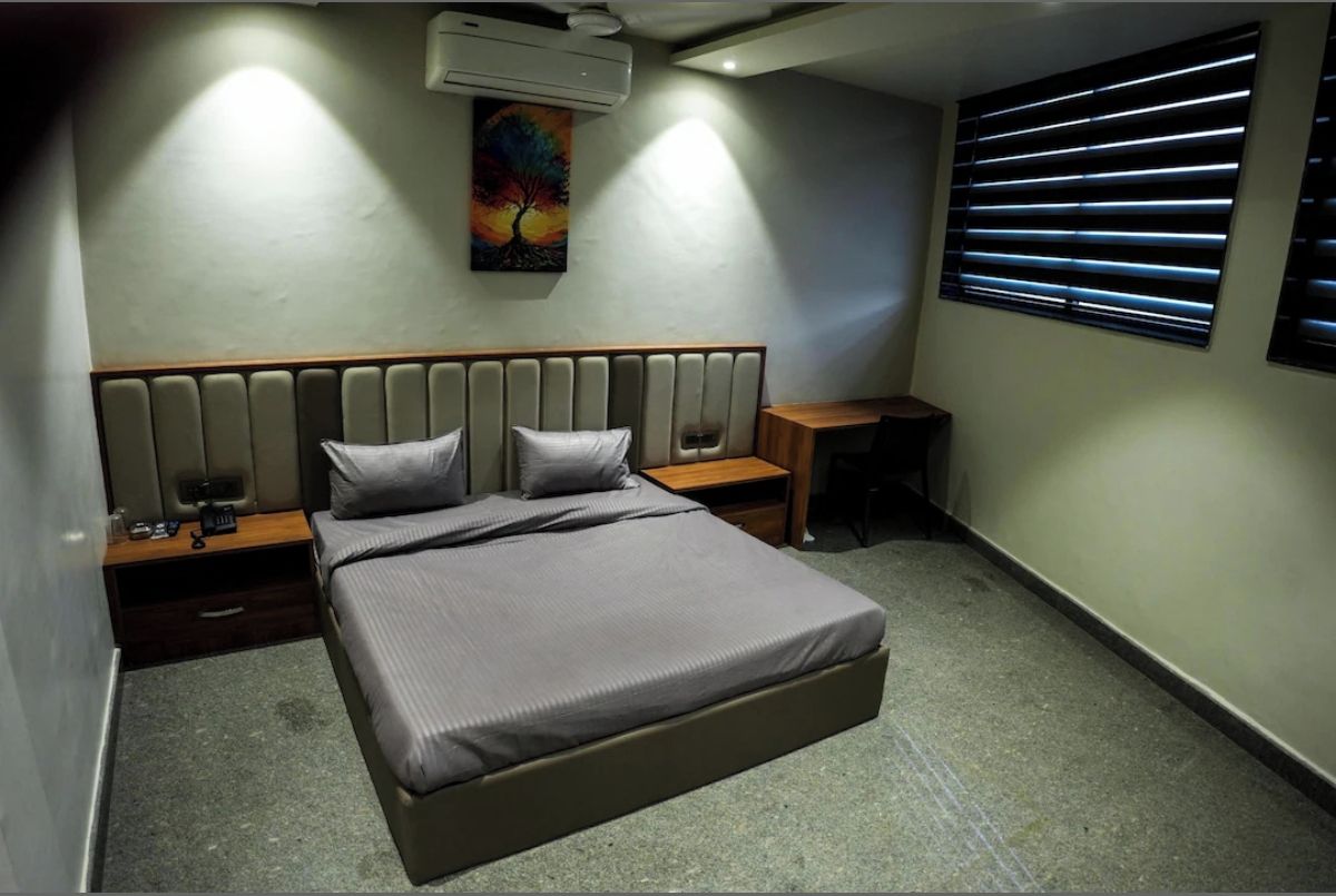 Un dormitorio con cama, mesitas de noche, aire acondicionado, un cuadro y una ventana con persianas.