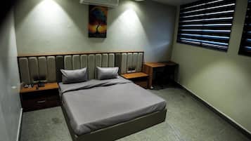 Un dormitorio con cama, mesitas de noche, aire acondicionado, un cuadro y una ventana con persianas.