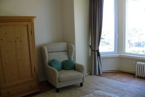 A room with a wooden cabinet, a beige armchair with green cushions, a window with curtains, and a radiator. - Brittany / Locquirec (Locquirec)