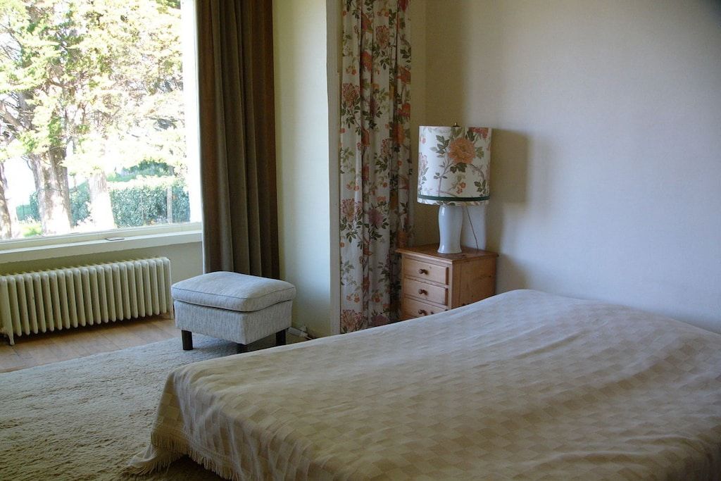 A bedroom with a bed, a nightstand, a lamp, a window with curtains, and a radiator.