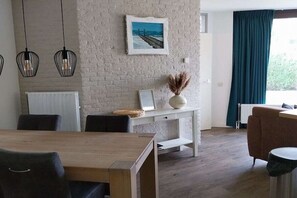 A modern living room with a wooden dining table, a white console table with decorative items, a sofa, and a view of the outdoors through a window with curtains. - Holiday home in Brouwershaven with barbecue (Brouwershaven)