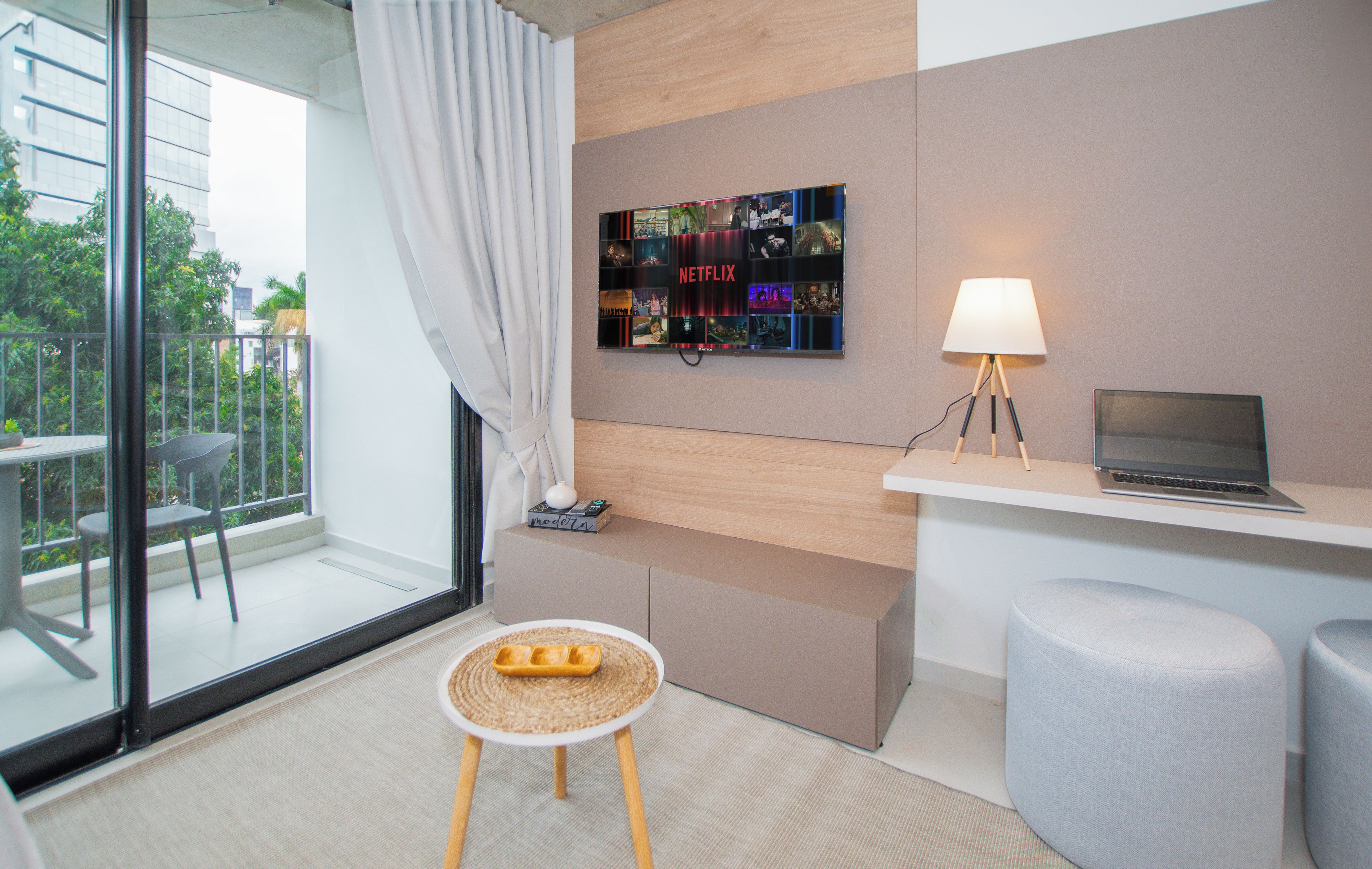 A modern hotel room with a glass door leading to a balcony, a wall-mounted Netflix display, a laptop on a desk, a small stool, and a lamp.