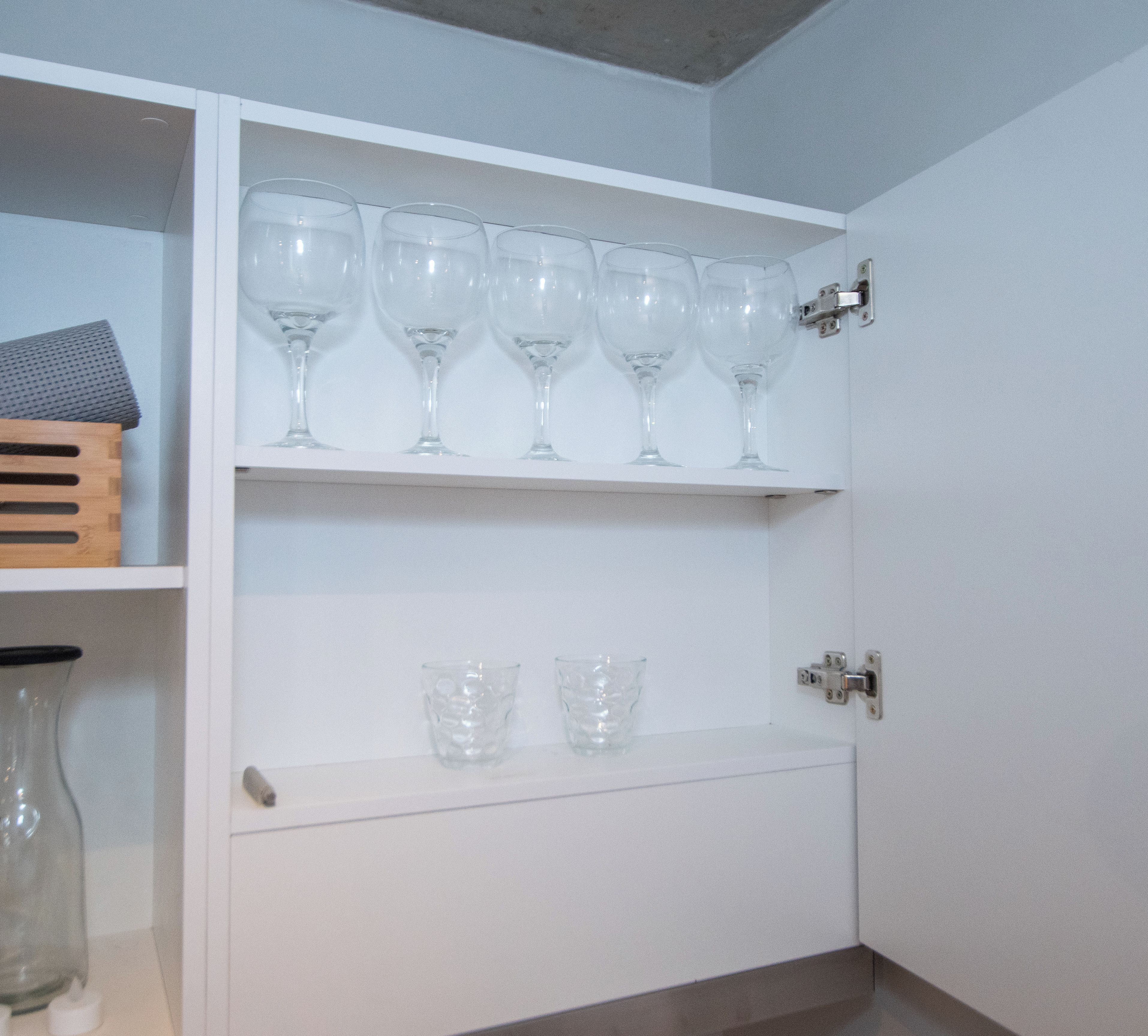Ein weißer Schrank mit Regalen, auf denen Weingläser und eine kleine Holz-Kiste stehen.