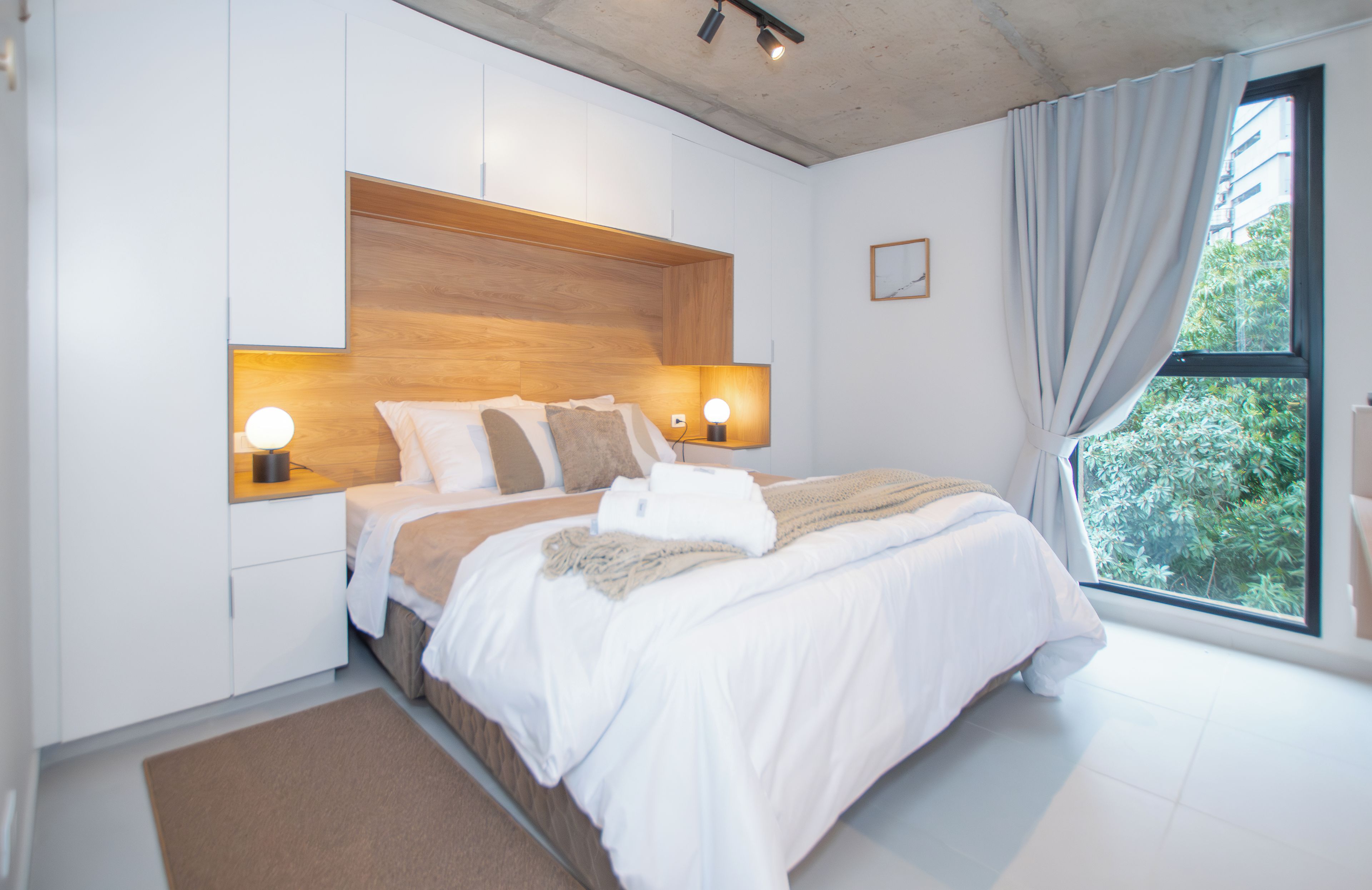 Ein modernes Schlafzimmer mit einem hölzernen Kopfteil, zwei Nachttischlampen, einem großen Bett mit weißer Bettwäsche und Blick auf Grün durch ein Fenster.