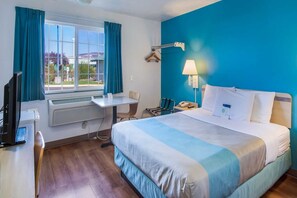 Habitació d'hotel amb un llit, un escriptori, una cadira, un televisor i una finestra amb cortines.