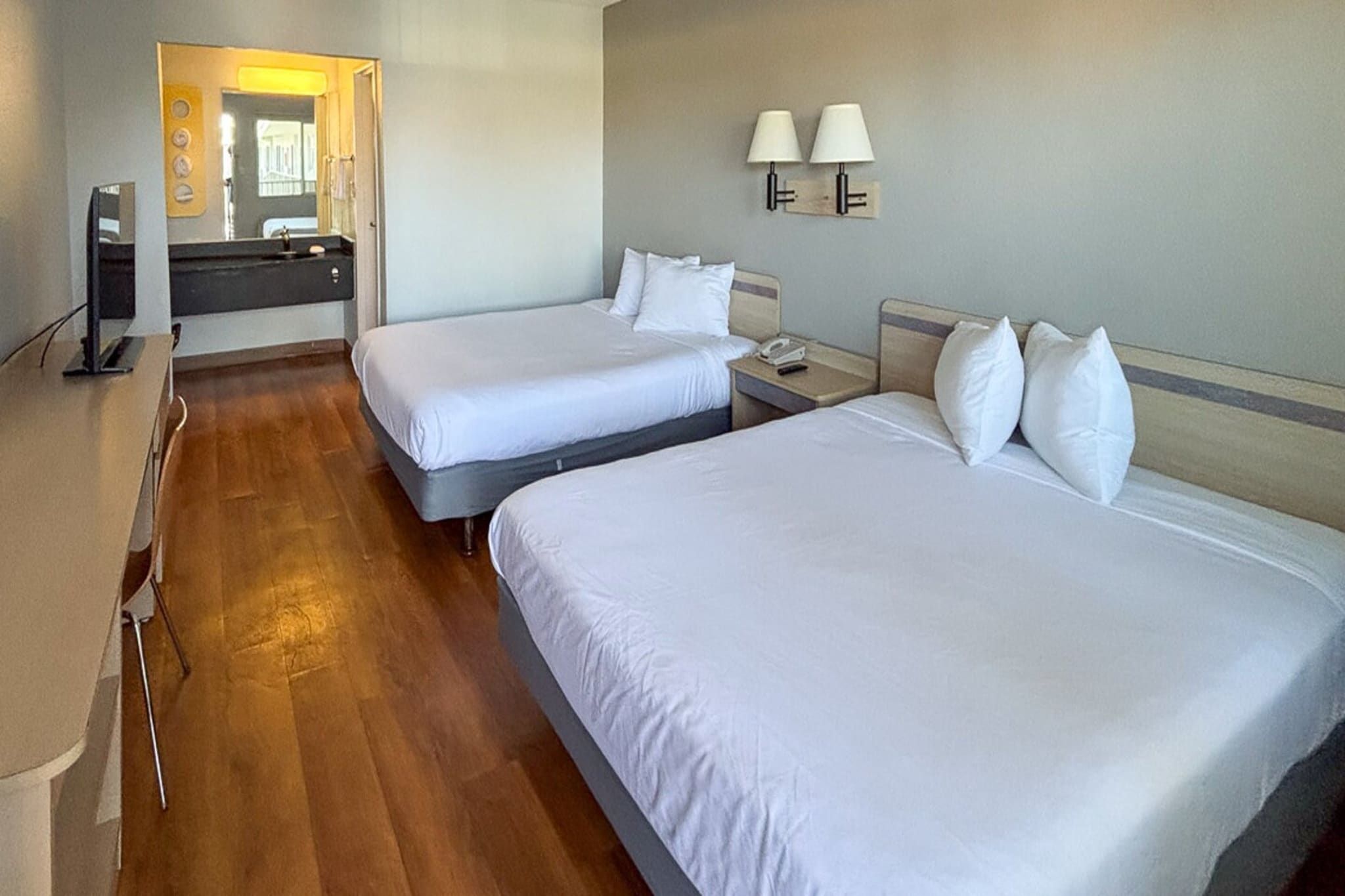 A hotel room with two beds, a bathroom with a mirror, and a desk with a lamp.