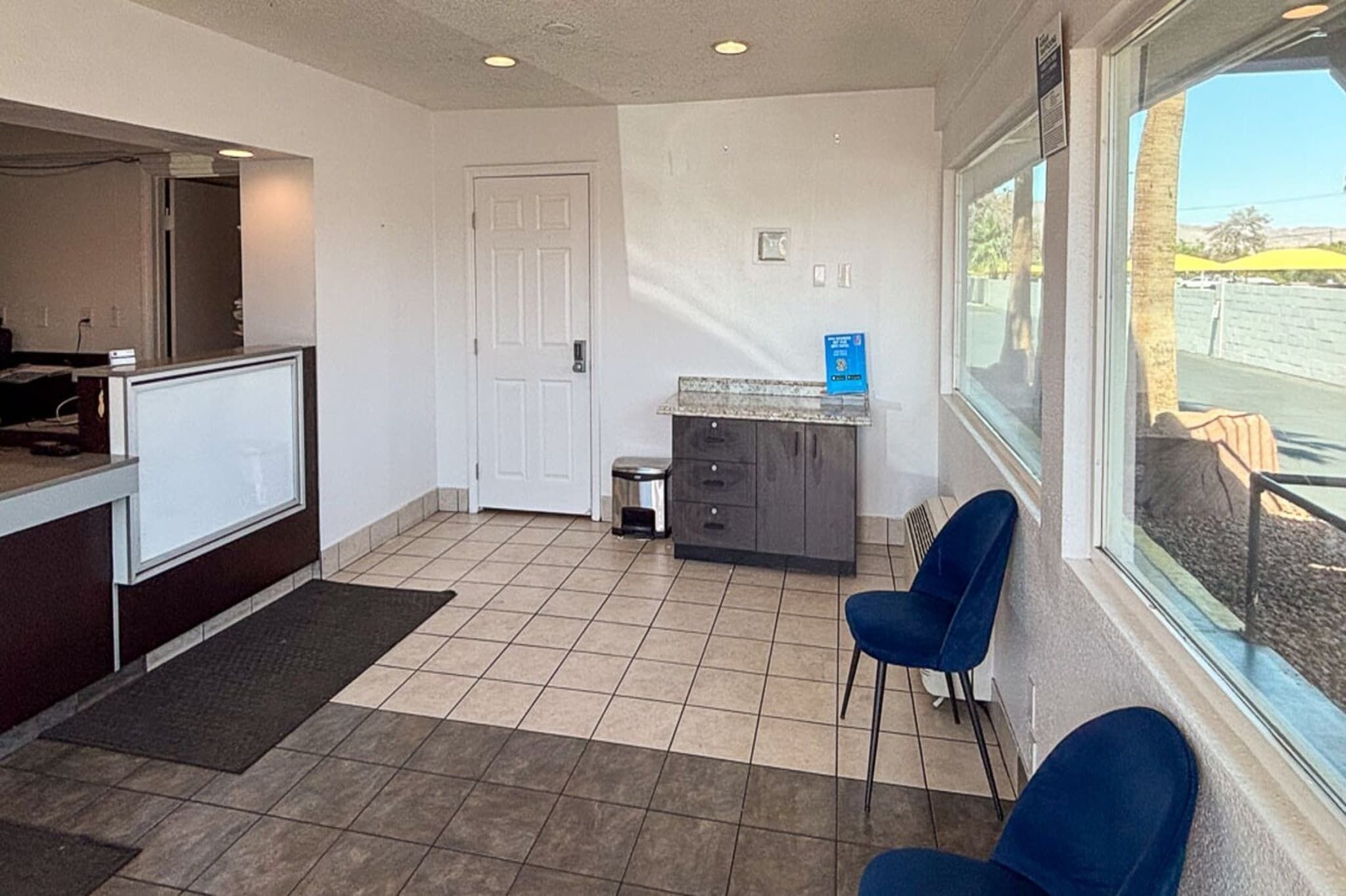 A reception area with a counter, a door, a small trash bin, a window with a view, and two blue chairs.
