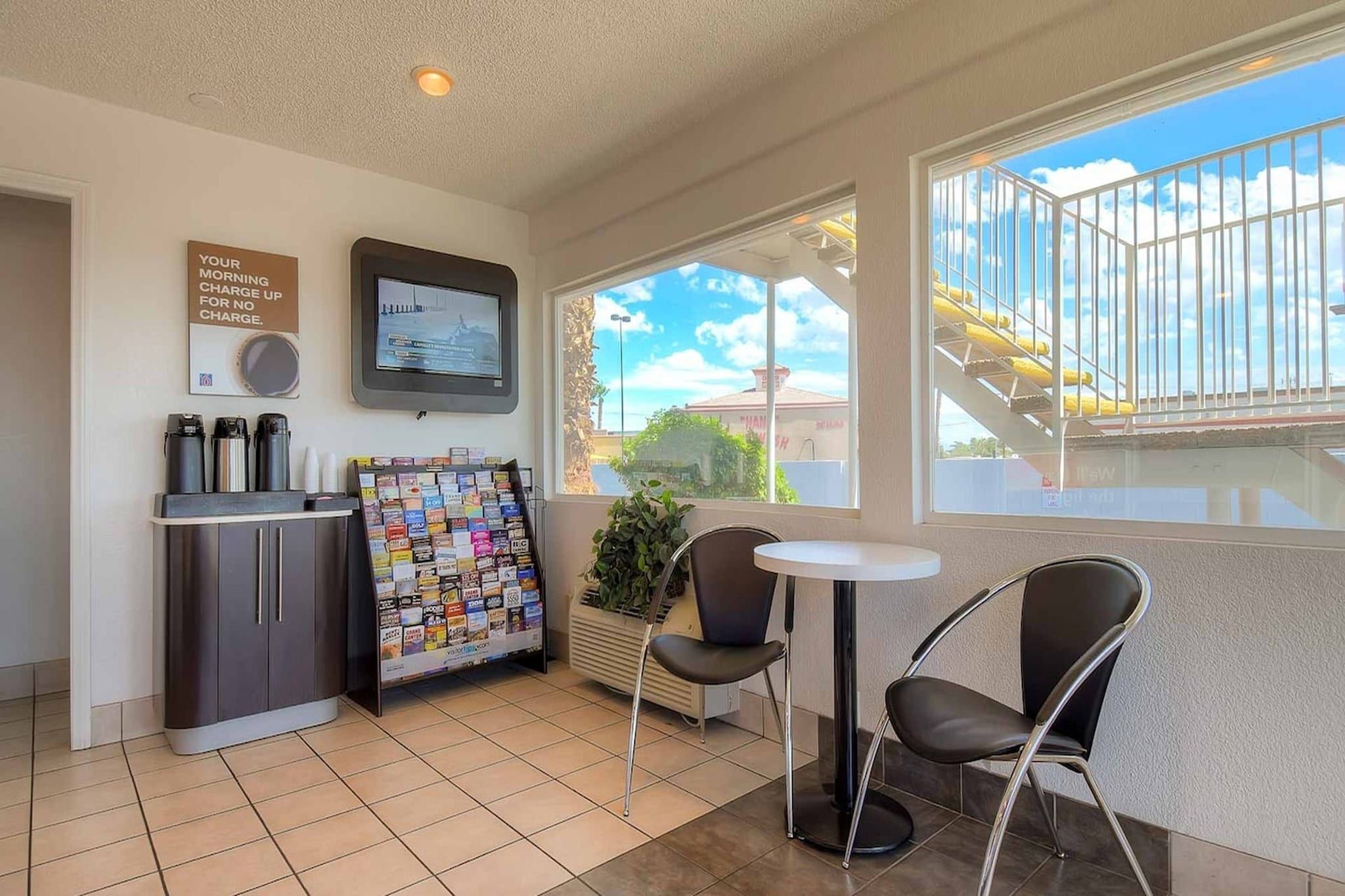 A small room with a kitchenette, a TV, a bulletin board, and a window with a view of a building and a staircase.