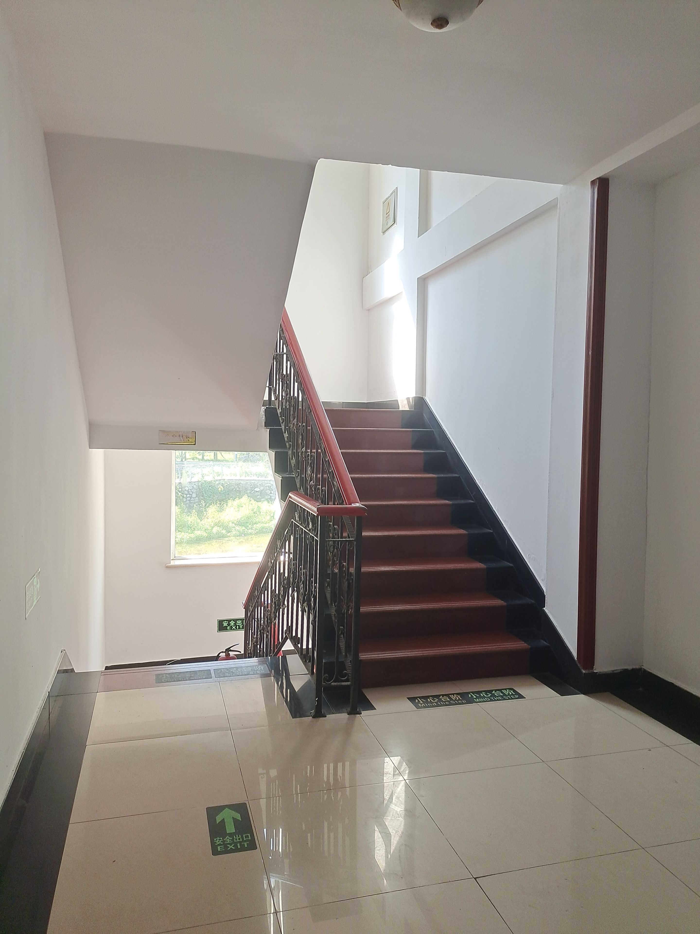 A staircase with a red handrail leading to an upper floor, a window with a view of greenery, and a tiled floor with directional arrows.