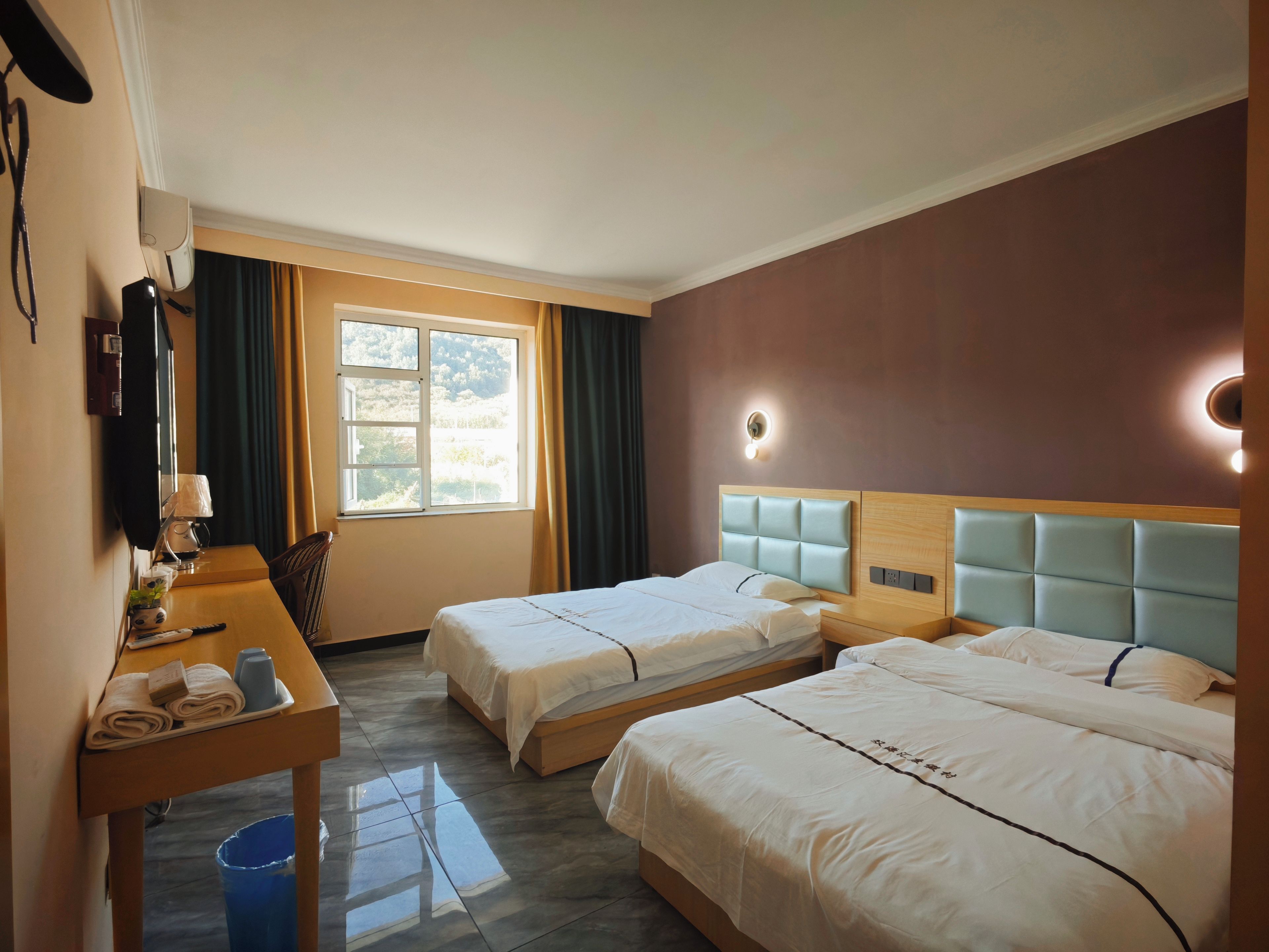 Ein Hotelzimmer mit zwei Betten, einem Schreibtisch mit Lampe, einem Fernseher und einem Fenster mit Vorhängen.