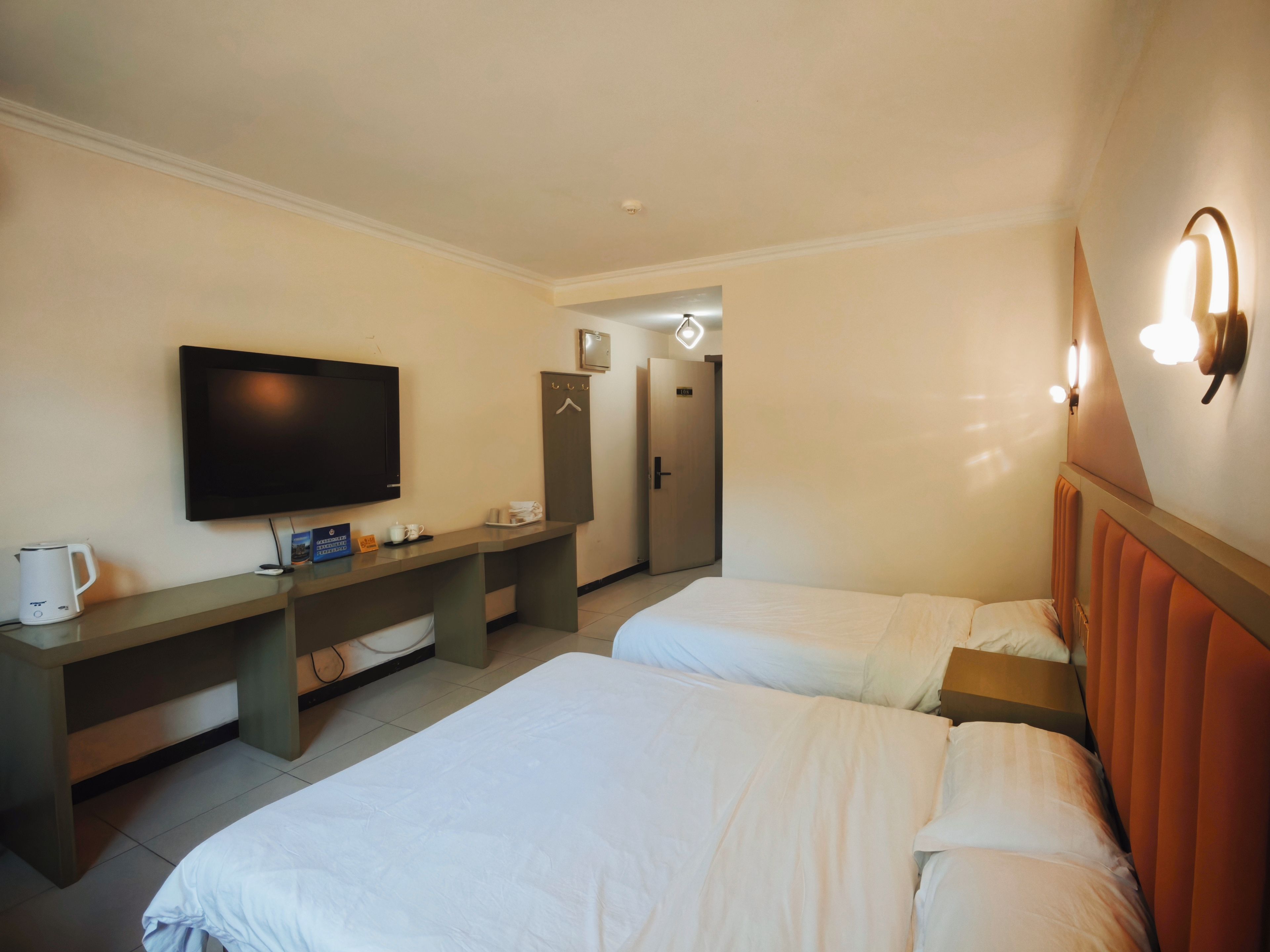 A hotel room with two beds, a television, a desk with a kettle, and a door leading to a bathroom.