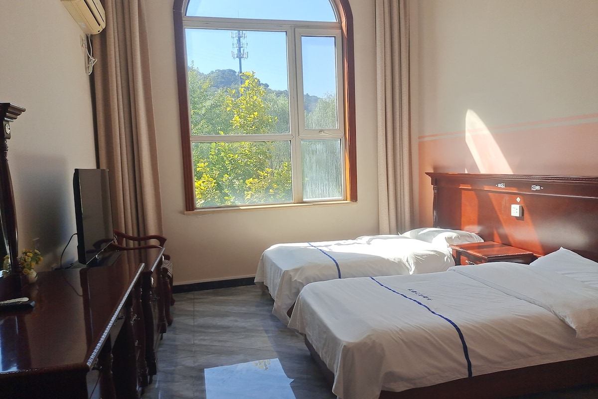 A hotel room with two beds, a desk, a chair, a TV, and a window with a view of trees and a power line.