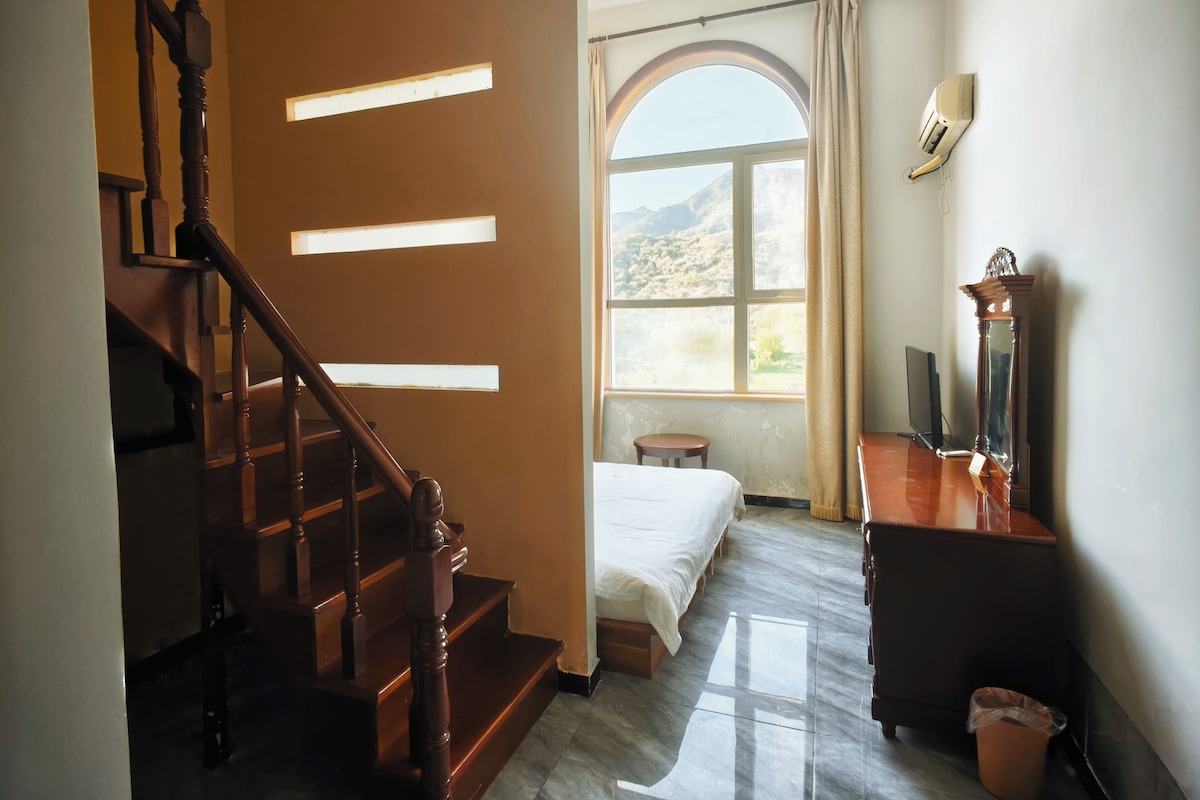 A room with a bed, a desk, a chair, a small stool, a window with a view of the outdoors, and a staircase.