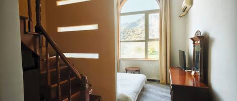 A room with a bed, a desk, a chair, a small stool, a window with a view of the outdoors, and a staircase.