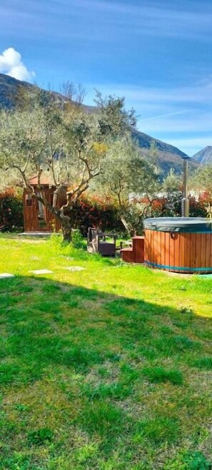 A wooden cabin with a hot tub and a small shed in a grassy yard with trees and mountains in the background.