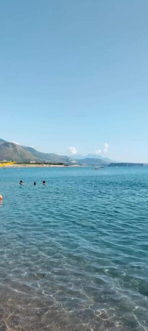 A clear view of a beach with calm water, distant mountains, and a few people swimming.