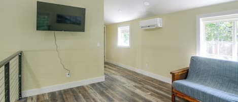 A room with a wooden floor, a blue sofa, a mounted TV, and an air conditioning unit.