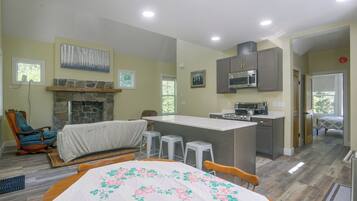 A spacious living area with a fireplace, a dining table with a floral tablecloth, a kitchen with modern appliances, and a view of the outdoors through windows.