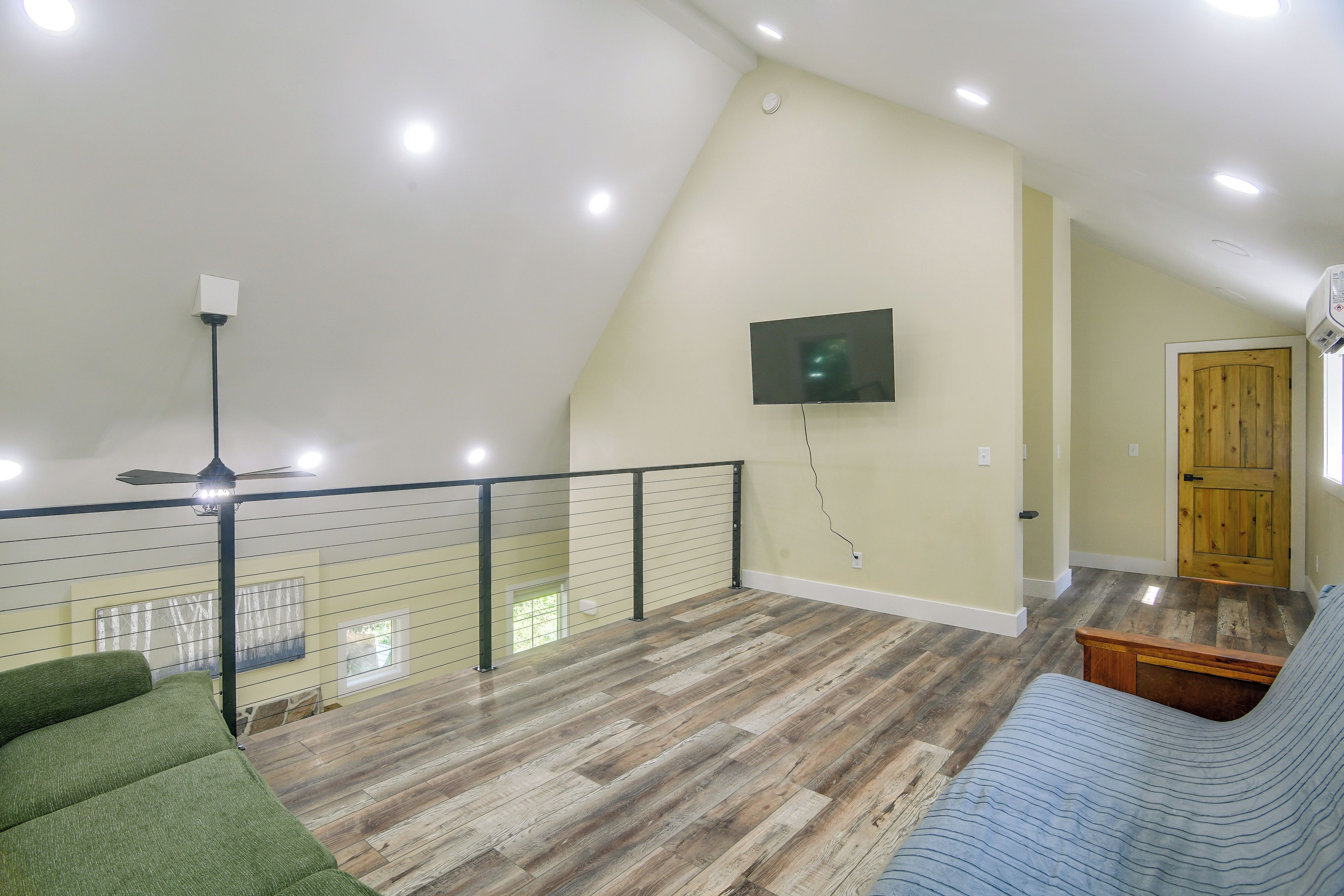 A spacious living area with a high ceiling, a flat-screen TV mounted on the wall, a wooden floor, and a green sofa.