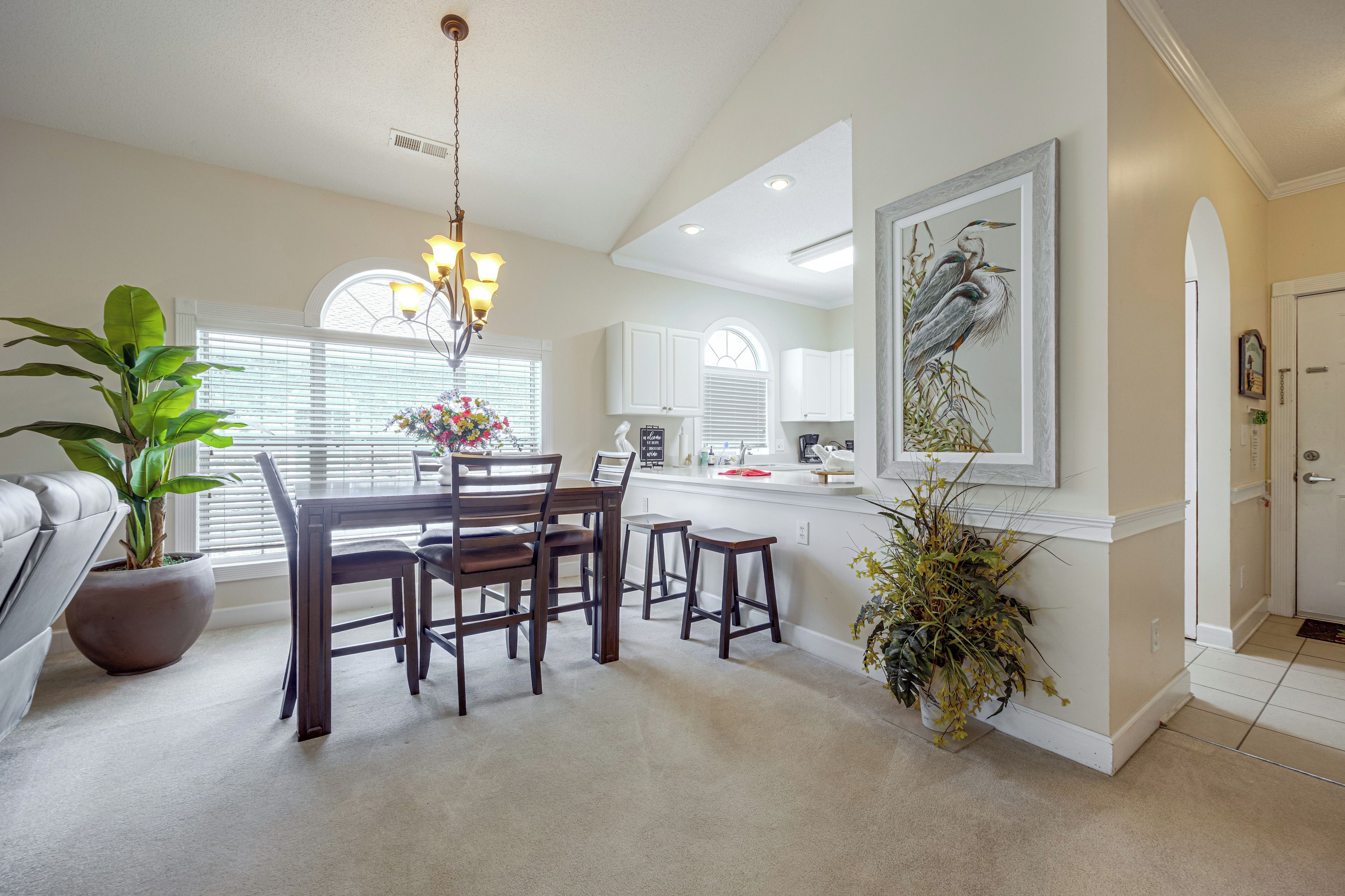 A modern kitchen with a dining area, featuring a chandelier, potted plants, and a large wall-mounted artwork.