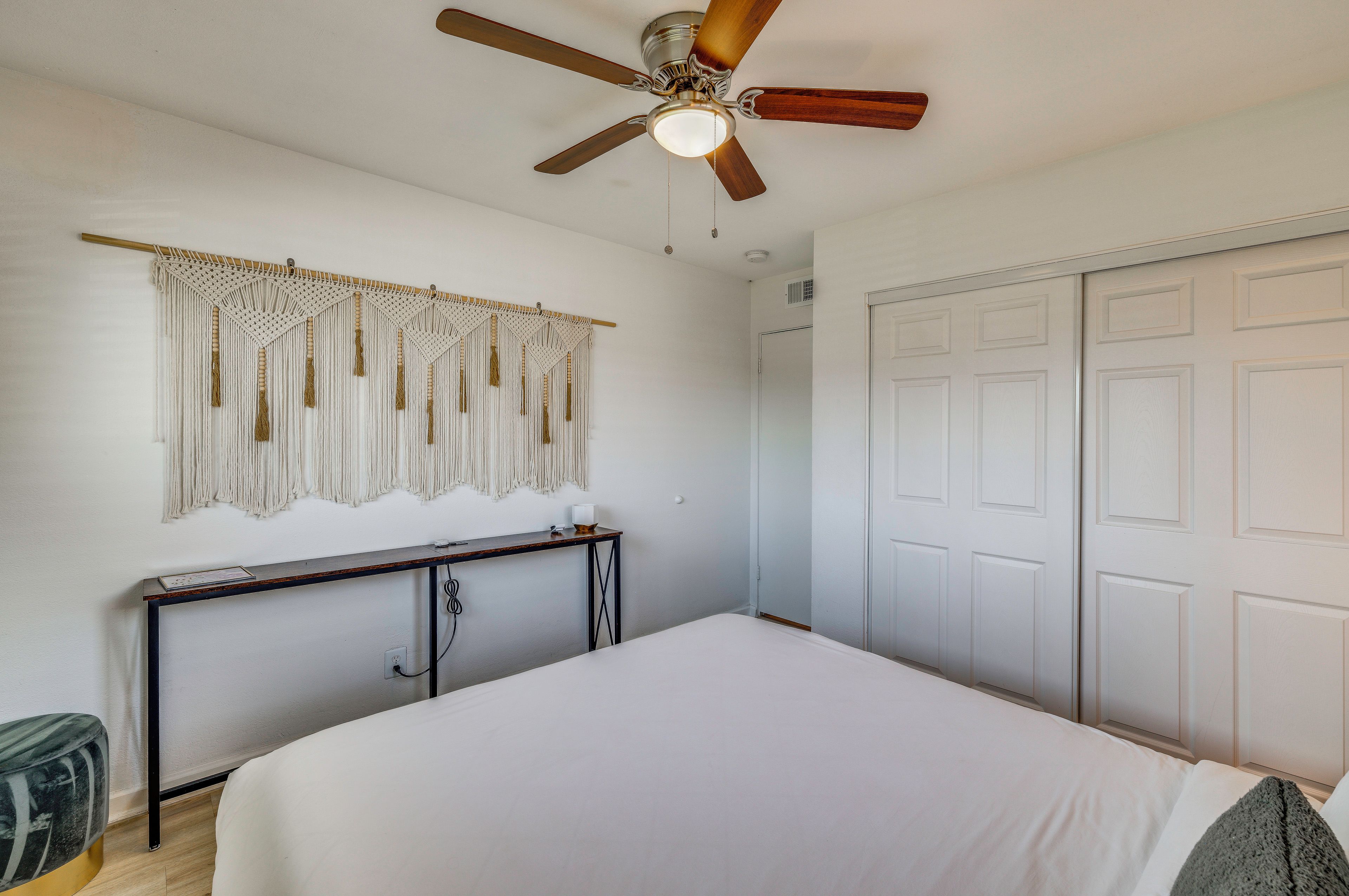 A bedroom with a bed, a ceiling fan, a wall-mounted macrame decoration, a bedside table, and a wardrobe.