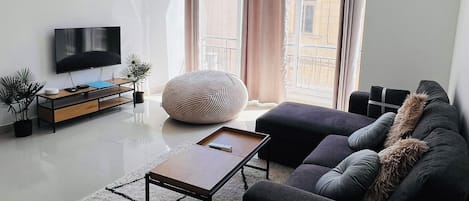 Una sala de estar moderna con una televisión de pantalla plana, un sofá, una mesa de centro y un ventanal con cortinas.
