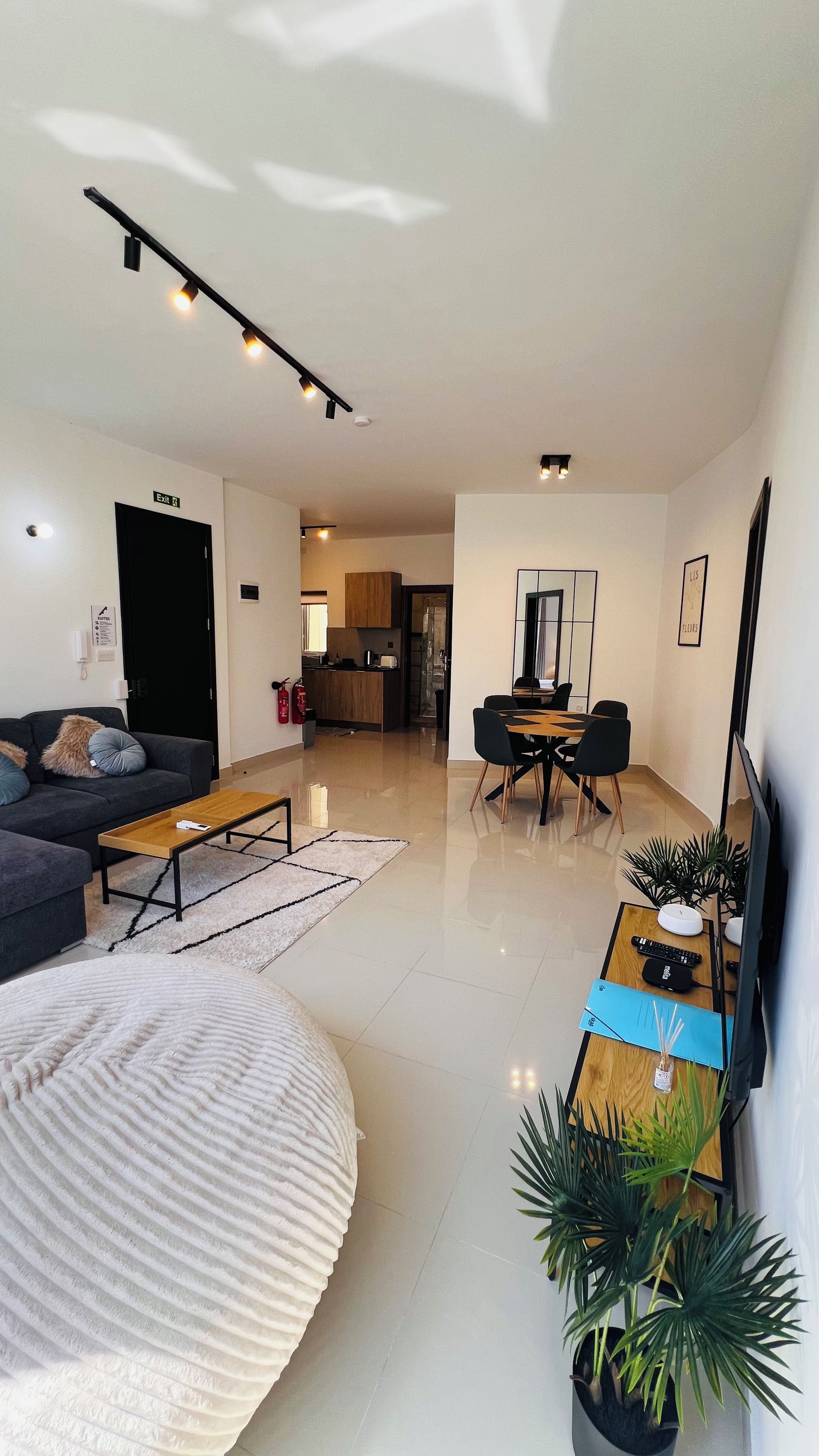 Una sala de estar moderna con sofá, mesa de centro y comedor con sillas y mesa. Se observa una cocina al fondo, un extintor en la pared y un televisor de pantalla plana montado en la pared.