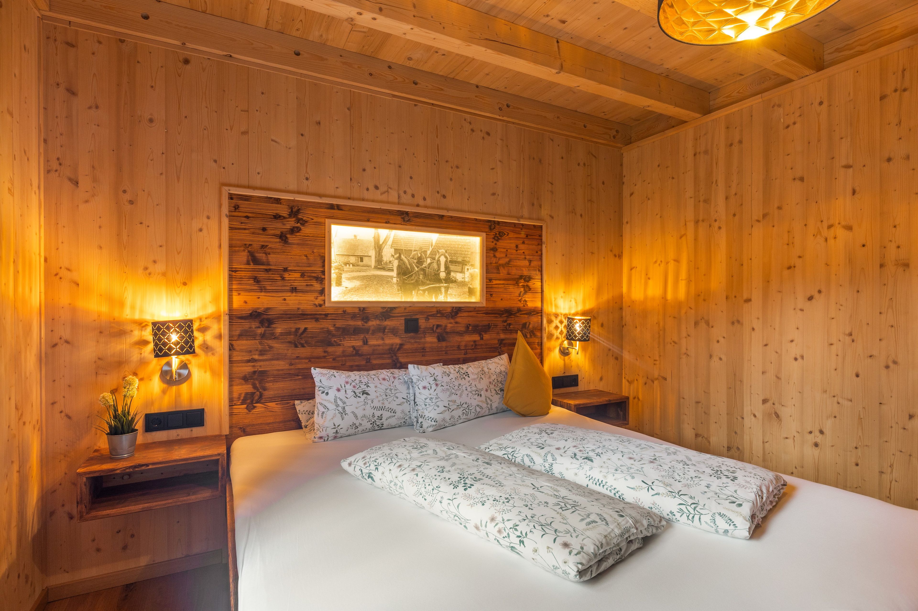 Een houten kamer met een bed, twee nachtkastjes, een aan de muur hangend schilderij en twee wandlampen.