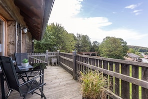 Une terrasse en bois avec une table et des chaises, d’où l’on a vue sur une zone boisée et sur des bâtiments situés au loin.