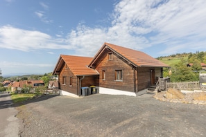 Une maison en bois à deux étages, avec un toit en pente, une allée gravillonnée et une vue sur une colline verdoyante.