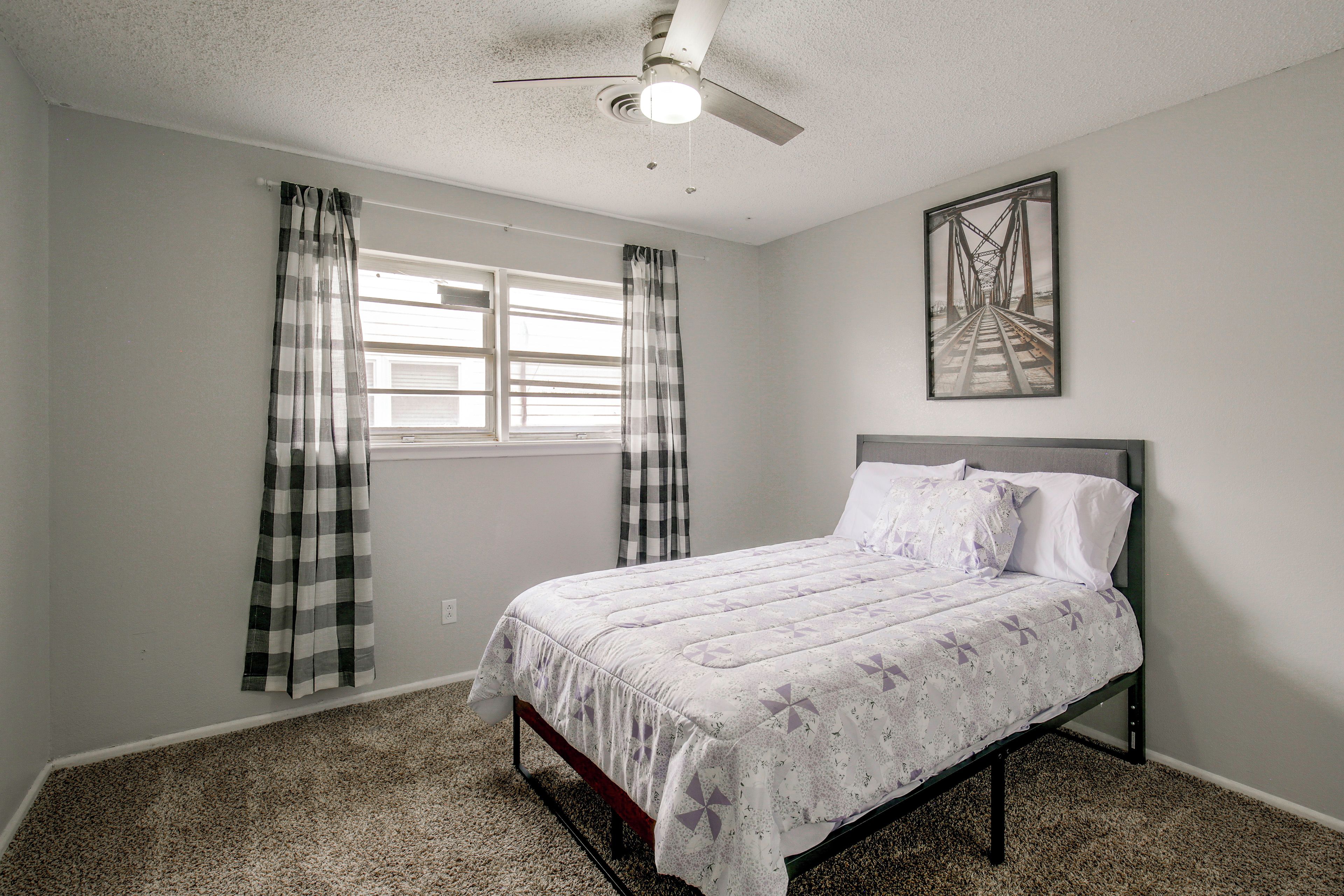 A bedroom with a bed, a ceiling fan, a window with curtains, and a wall-mounted picture.