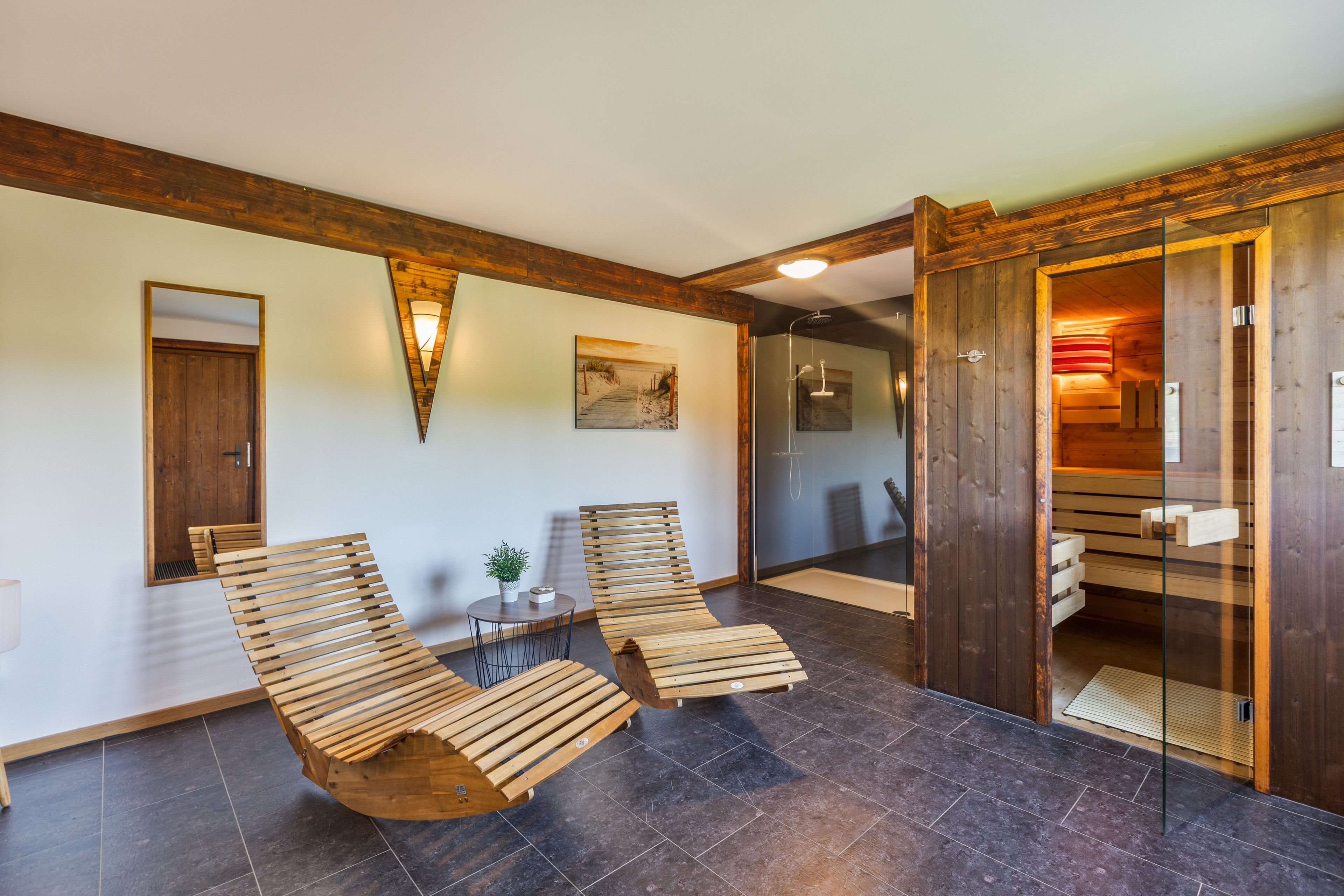 A room with two wooden lounge chairs, a small table with a plant, and a glass shower enclosure.