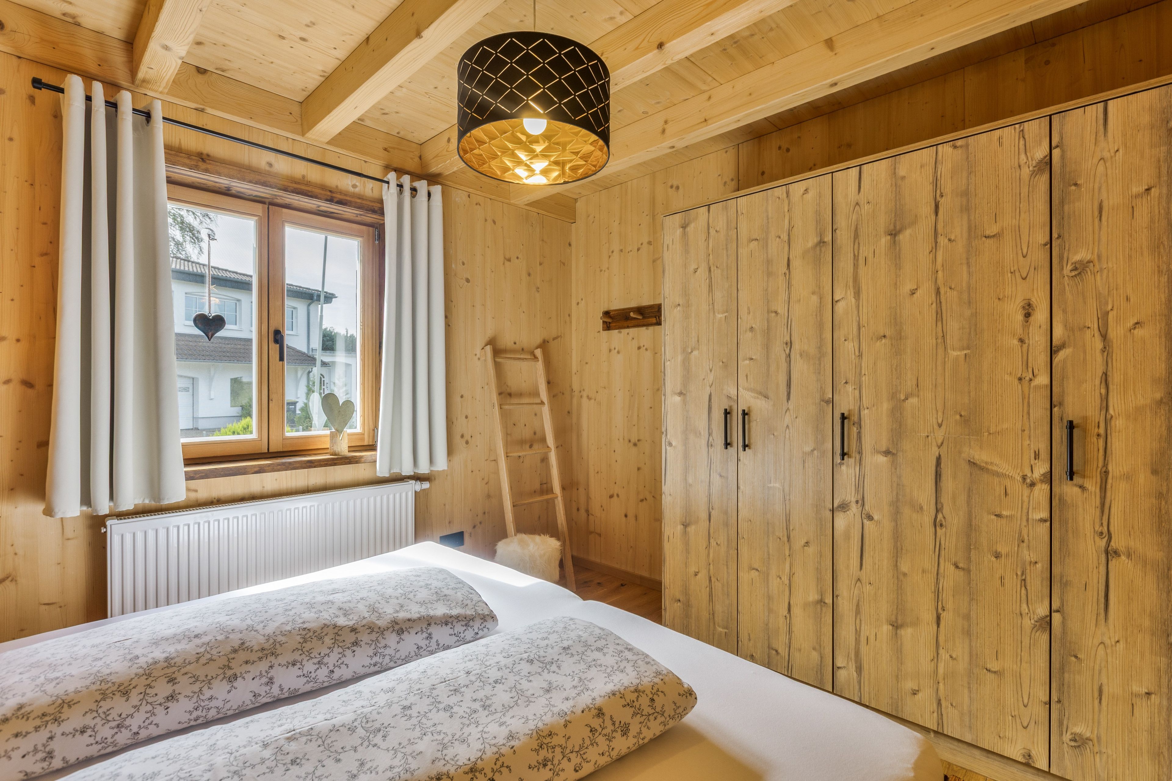 Een slaapkamer met houten vloer, een bed, een raam met gordijnen, een radiator en een kledingkast.
