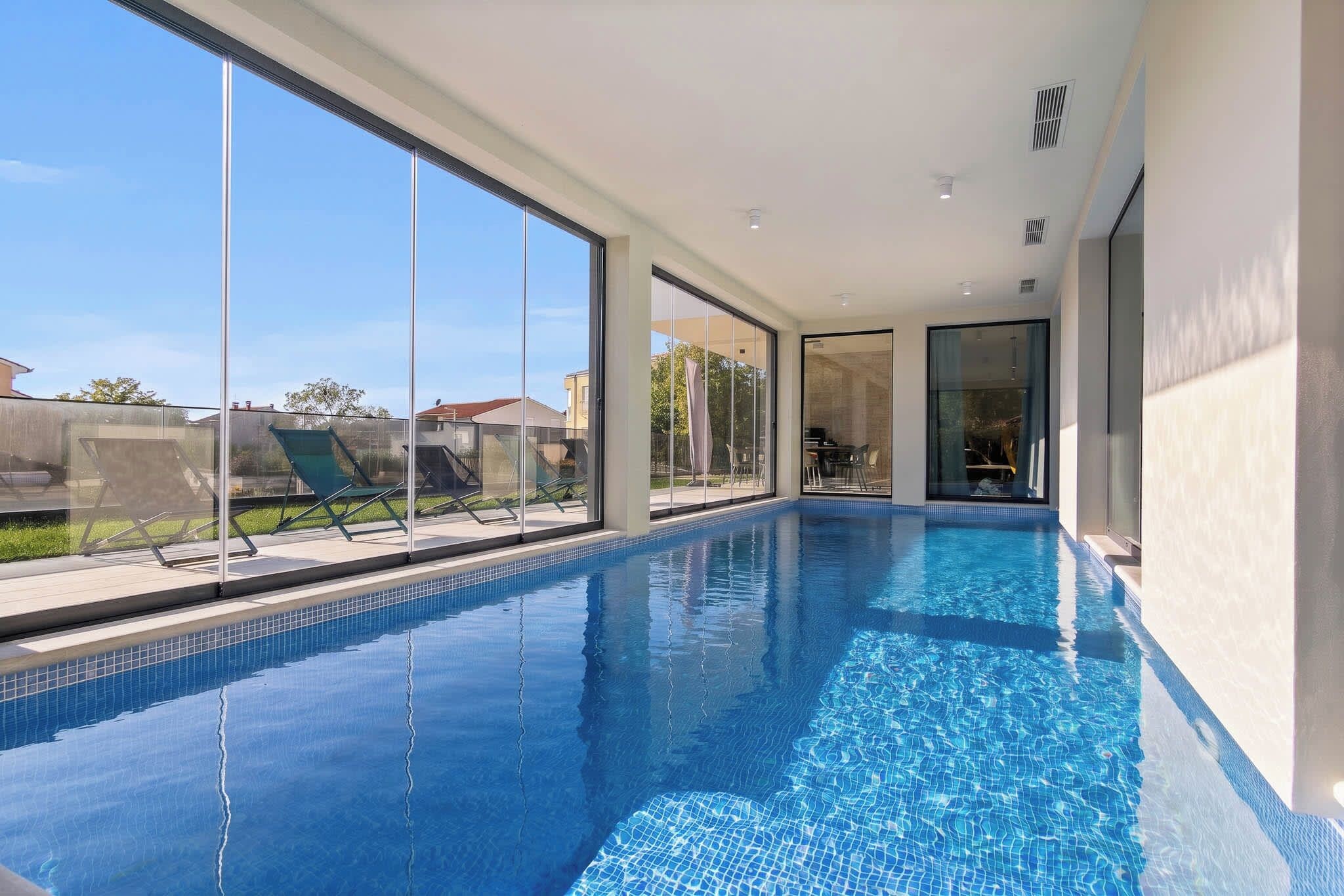 A modern indoor swimming pool with clear glass walls, a tiled floor, and a view of the outside.