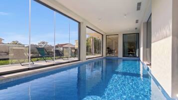 A modern indoor swimming pool with clear glass walls, a tiled floor, and a view of the outside.