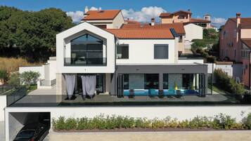 A modern two-story house with a flat roof, large windows, and a covered outdoor seating area.