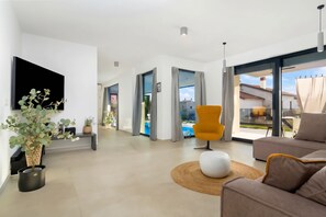 A modern living room with a flat-screen TV, a sofa, a yellow chair, and a large window offering a view of a swimming pool and buildings.