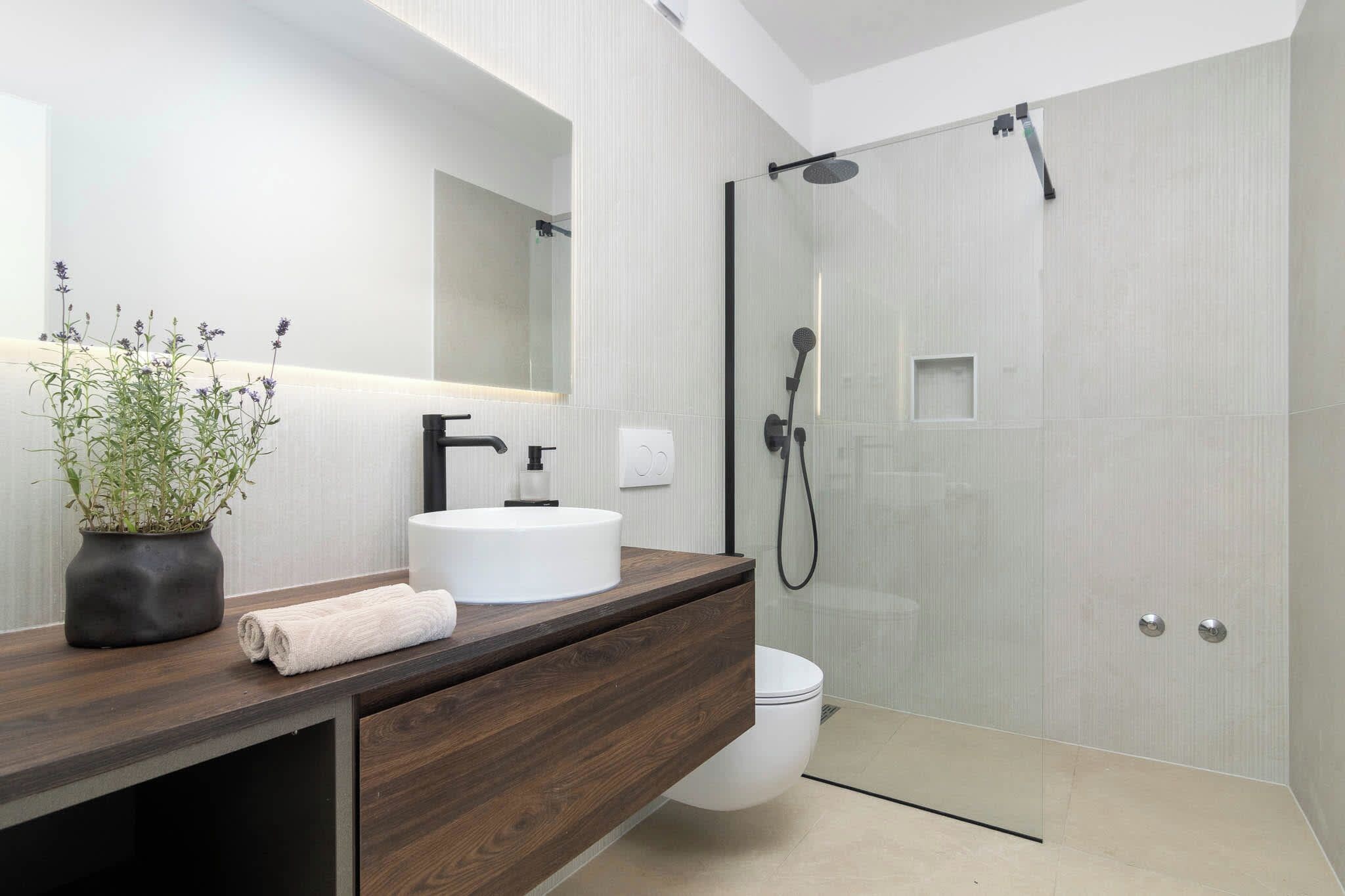 A modern bathroom with a wooden vanity, a white sink, a glass shower enclosure, and a potted plant.